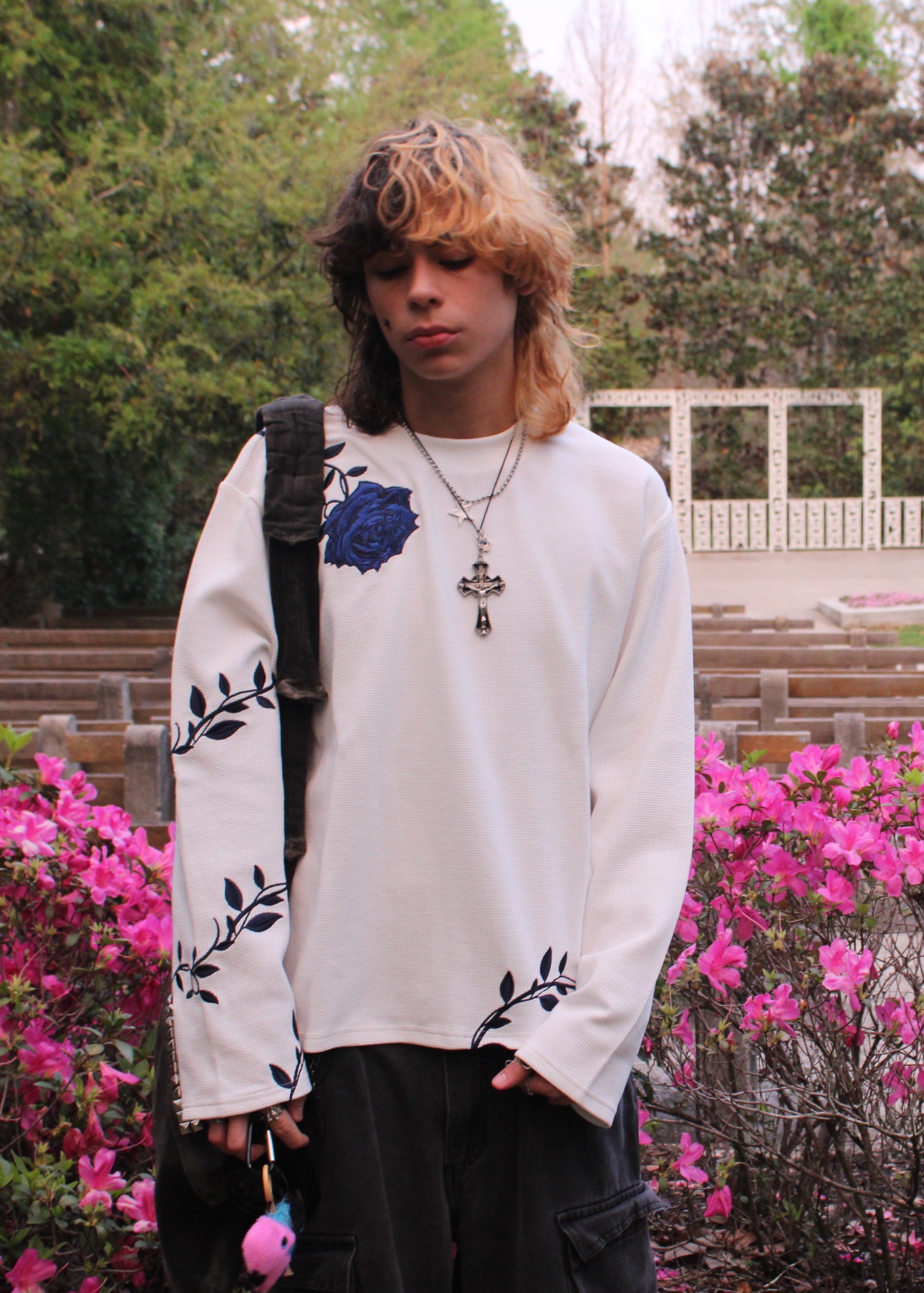 Person wearing a white long-sleeve shirt with floral designs, standing in a park with pink flowers and green trees.
