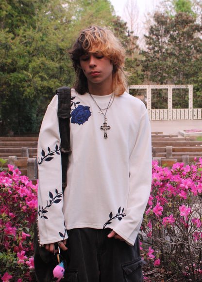 Person wearing a white long-sleeve shirt with floral designs, standing in a park with pink flowers and green trees.